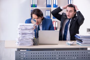 Two young men in business suits are sitting at a desk in an office. One is holding a magnifying glass up to a tall stack of papers and the other is holding his hands up to his head and expressing overwhelm.