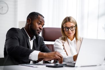 Two people looking at a laptop screen while sitting at a table in the office.