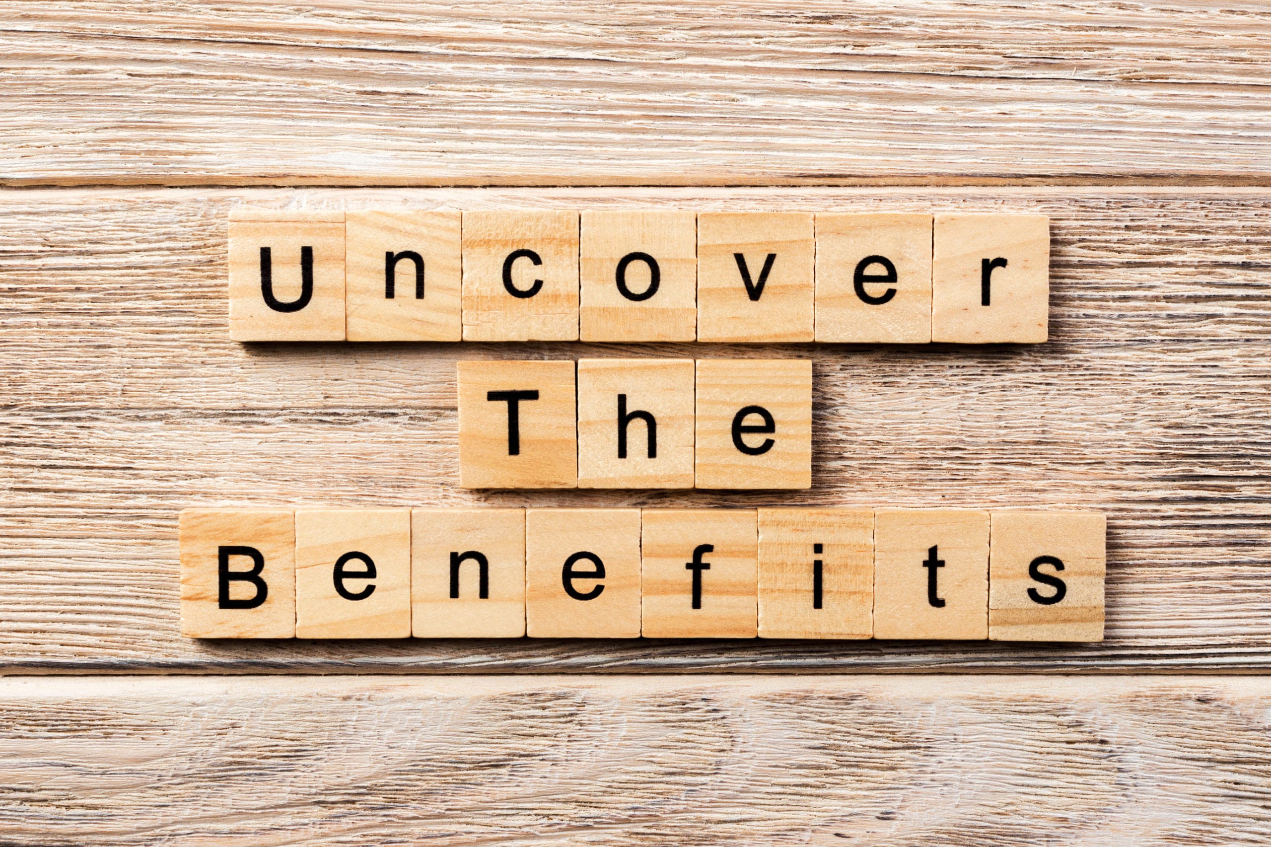 The words "Uncover The Benefits" on wooden letter blocks against a wooden background.