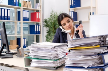 A business person sitting at a desk talking on two different phones surrounded by piles of loose papers in an office.