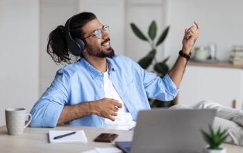 A person sitting a desk with laptop. The person is smiling and playing air guitar.