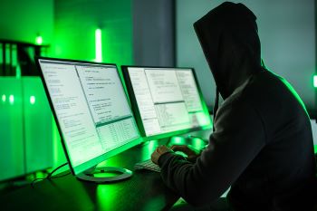 A person sitting in a dimly lit room in front of two computer monitors while wearing a dark hoodie with the hood up.