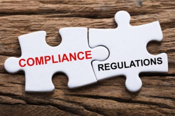 Two white puzzle pieces on a wood background. One puzzle pieces has the word "Compliance" on it and one has the word "Regulations" on it.