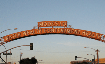 A large red sign that arcs overtop of a street that reads "Modesto Water Wealth Contentment Health"